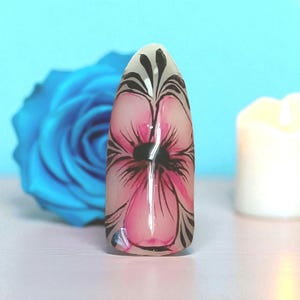 Puede incluir: Una sola uña acrílica con un diseño floral. La uña presenta una flor rosa con contornos negros y un centro negro. La uña está sobre un fondo azul claro con una rosa azul y una vela encendida.