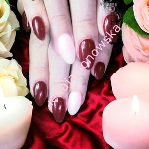 Puede incluir: Primer plano de uñas manicuradas en una variedad de colores. Algunas uñas son de un borgoña profundo, mientras que otras son de un rosa suave. Las uñas son de forma ovalada y brillantes. El fondo incluye velas y flores.