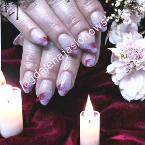Può includere: Unghie curate con base trasparente e un design con sfumature di viola chiaro e rosso intenso. Le unghie sono lunghe e di forma ovale. L'immagine include anche candele accese e fiori.