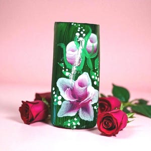 Puede incluir: Un jarrón cilíndrico verde pintado a mano con rosas rosas y blancas y tallos verdes. Puntos blancos acentúan el diseño floral. Varias rosas rojas están dispuestas alrededor de la base del jarrón sobre una superficie rosa.