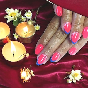 Op de afbeelding: Felroze nagellak met blauwe uiteinden en witte accenten. De nagels bevinden zich op een hand, tegen een rode satijnen achtergrond met brandende kaarsen en kleine witte en gele bloemen.
