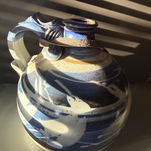 Op de afbeelding: Een handgemaakte keramische kruik met een afgeronde body en een opvallende handgreep. De kruik heeft een blauw-wit abstract ontwerp, met een donkerblauwe rand en handgreep. De kruik is een decoratief stuk.