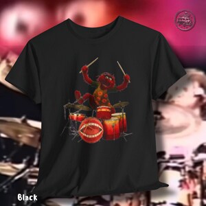 K&ouml;nnte beinhalten: Schwarzes T-Shirt mit einem Cartoon-Schlagzeuger. Die Figur ist rot mit gelben und orangen Details und h&auml;lt Trommelst&ouml;cke. Das Schlagzeug ist rot und goldfarben. Das Wort "Black" ist unten auf dem Shirt aufgedruckt.