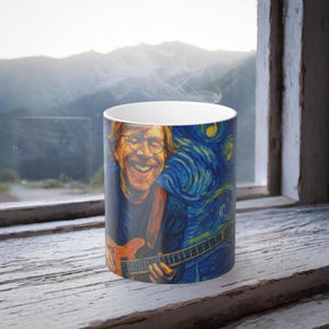 May include: A white ceramic mug featuring a smiling man playing a guitar, set against a swirling blue and yellow starry night background. The mug has a white interior and is likely filled with a hot beverage, as steam rises from the top.