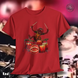 K&ouml;nnte beinhalten: Ein rotes T-Shirt mit einer Cartoon-Figur, die Schlagzeug spielt. Die Figur ist rot mit gelben und orangefarbenen Akzenten. Das Schlagzeug ist rot und orange. Das Wort "Red" ist unten links in Wei&szlig; gedruckt.