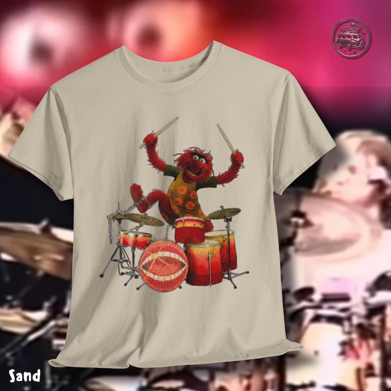 K&ouml;nnte beinhalten: Sandfarbenes T-Shirt mit einer Zeichentrickfigur, die ein Schlagzeug spielt. Die Figur ist rot mit einem gr&uuml;nen Hemd und gelbem Blumenmuster. Das Schlagzeug ist rot und goldfarben. Das Wort "Sand" ist unten links aufgedruckt.