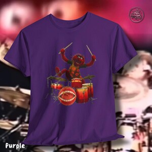 K&ouml;nnte beinhalten: Lila T-Shirt mit einer Zeichentrickfigur, die ein Schlagzeug spielt. Die Figur ist rot mit einem gelb-orange gemusterten Hemd. Das Schlagzeug ist rot und orange. Das Wort "Purple" steht unten auf dem T-Shirt.