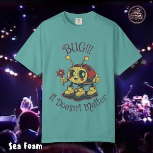 May include: Sea foam green t-shirt featuring a cartoon ladybug holding a flower and giving a thumbs up. The text "BUG!!! It Doesn't matter" is printed above and below the ladybug illustration. The shirt has a crew neck.