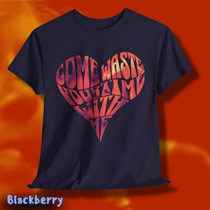 May include: A navy blue t-shirt featuring a heart-shaped design with the words "COME WASTE YOUR TIME WITH ME" in a gradient of orange, red, and purple. The shirt has short sleeves and a crew neck.