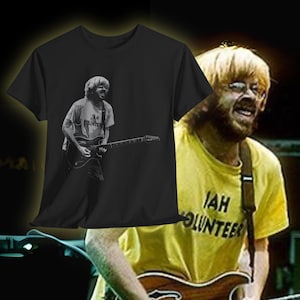 May include: Black t-shirt featuring a grayscale image of a musician playing a guitar. The musician is wearing a shirt that says "JAH VOLUNTEER". The image is set against a yellow shirt with the same text.