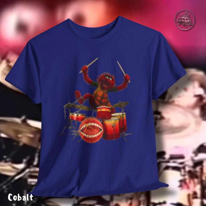 K&ouml;nnte beinhalten: Kobaltblaues T-Shirt mit einem Cartoon-Schlagzeuger in Rot und Gelb. Das Schlagzeug ist rot und goldfarben. Das Wort "Cobalt" ist unten auf dem Shirt aufgedruckt. Der Hintergrund ist verschwommen.