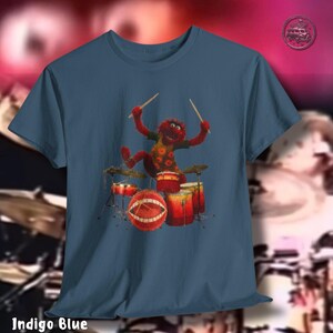 K&ouml;nnte beinhalten: Indigoblaues T-Shirt mit einer Zeichentrickfigur, die Schlagzeug spielt. Die Figur ist rot mit einem gelb-orange gemusterten Hemd. Das Schlagzeug ist rot und orange. Der Text "Indigo Blue" befindet sich am unteren Rand des T-Shirts.