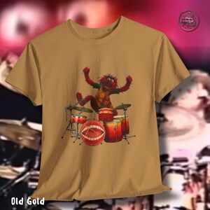 K&ouml;nnte beinhalten: Oltgoldfarbenes T-Shirt mit einem Cartoon-Schlagzeuger. Die Figur ist rot mit einem gr&uuml;nen Hemd und h&auml;lt Trommelst&ouml;cke. Das Schlagzeugset umfasst eine rote Bassdrum mit Munddesign und andere rote Trommeln.