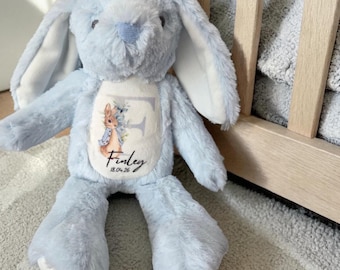 May include: A light blue plush bunny rabbit with long ears and a white belly. The bunny has a personalized design on its chest with the name "Finley" and the date "18.04.26". The bunny is sitting on a light gray rug.