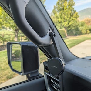 Puede incluir: Un soporte para teléfono LISEN negro está fijado al salpicadero de un vehículo. El soporte está situado cerca de la rejilla de ventilación y está conectado a un brazo de soporte. El interior del coche es visible, incluyendo el espejo retrovisor y una agarradera.