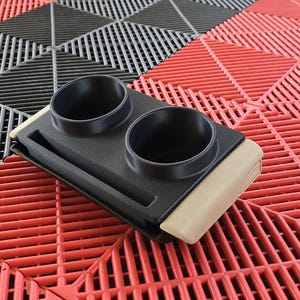 May include: A black plastic cup holder with two circular openings and a small storage area. The cup holder has a tan-colored base and is set on a red, black, and gray interlocking floor tile.