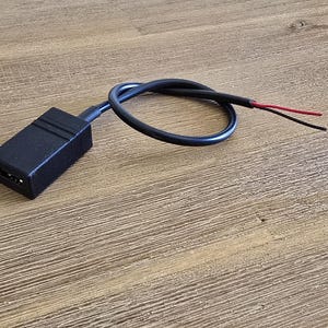 May include: A black electrical connector with a black cable and exposed red and black wires. The connector is rectangular with two small holes. The cable is looped and lies on a wooden surface.