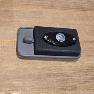 Puede incluir: Una carcasa rectangular de plástico gris con un inserto rectangular negro y una luz LED negra ovalada centrada. La lámpara contiene tres pequeñas bombillas circulares y está asegurada con dos tornillos plateados en los lados izquierdo y derecho.