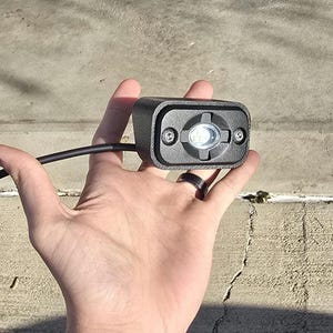 May include: A black LED light with a rectangular housing, held in a hand. The light has a clear lens and is secured with two screws. A black cable and connector extend from the light.