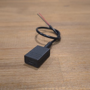 Könnte beinhalten: Ein schwarzes rechteckiges elektronisches Bauteil mit einem USB-Anschluss, verbunden mit einem schwarzen Kabel mit roten und weißen Drähten. Das Bauteil liegt auf einer Holzoberfläche.