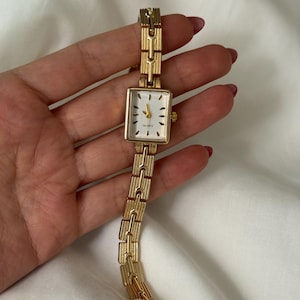 Puede incluir: Un reloj de cuarzo dorado con una esfera rectangular y un brazalete de eslabones a juego. La esfera del reloj es blanca con marcadores de hora y agujas negras. El reloj se sostiene en una mano.