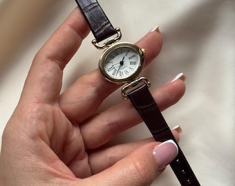 Reloj de pulsera vintage para mujer con correa de cuero negro, caja redonda dorada y esfera con números romanos. Movimiento de cuarzo.