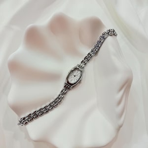 Könnte beinhalten: Eine silberfarbene Armbanduhr mit ovalem Zifferblatt und Gliederarmband. Die Uhr liegt auf einer weißen, muschelförmigen Dekoschale vor einem Hintergrund aus weißem Stoff. Das Zifferblatt der Uhr zeigt schwarze Ziffern und Zeiger.