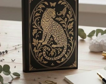 May include: Black journal with a gold leopard design. The cover features a detailed illustration of a leopard surrounded by ornate floral elements and a butterfly. The journal is standing upright on a wooden surface.