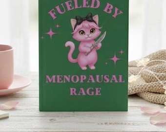 Puede incluir: Un libro de tapa dura verde oscuro con un gato de dibujos animados rosa que lleva un lazo negro y sostiene un cuchillo. El texto FUELED BY MENOPAUSAL RAGE está impreso en rosa sobre y bajo el gato, con pequeños gráficos de estrellas rosas.