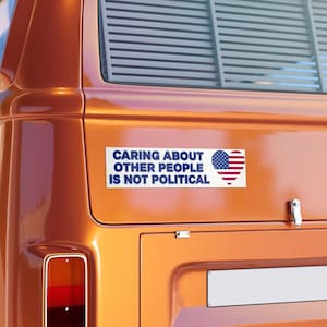 Könnte beinhalten: Ein rechteckiger Aufkleber mit dem Text "CARING ABOUT OTHER PEOPLE IS NOT POLITICAL" in blauen Buchstaben. Der Aufkleber zeigt auch ein herzförmiges Design mit der amerikanischen Flagge.