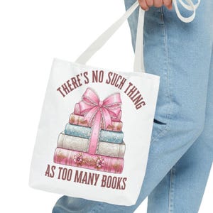 May include: White tote bag with a graphic of a stack of books tied with a pink bow. The text on the bag reads "THERE'S NO SUCH THING AS TOO MANY BOOKS" in brown lettering. The bag has white handles.