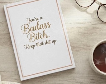 May include: A white notebook with a gold border and the text "You're a Badass Bitch. Keep that shit up." is displayed. The notebook is next to a pair of glasses and a cup of coffee.