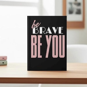 Op de afbeelding: Een zwart notitieboek met de tekst "be BRAVE BE YOU" in witte en roze letters. Het notitieboek ligt op een houten oppervlak. De achtergrond bevat een raam en een ingelijste foto.