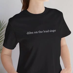 May include: Black t-shirt with the white text "dibs on the lead singer". The short-sleeved top has a crew neck. The text is centrally placed on the chest.