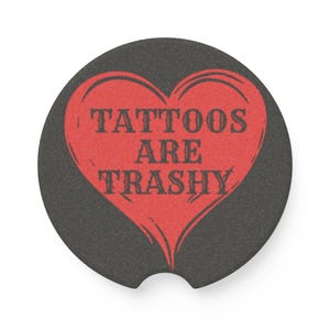 Puede incluir: Un posavasos negro para coche con un diseño de corazón rojo. El corazón contiene el texto "TATTOOS ARE TRASHY" en una fuente desgastada. El posavasos es redondo con una hendidura curva para facilitar su extracción.