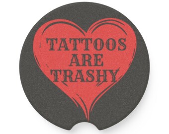 Puede incluir: Un posavasos negro para coche con un diseño de corazón rojo. El corazón contiene el texto "TATTOOS ARE TRASHY" en una fuente desgastada. El posavasos es redondo con una hendidura curva para facilitar su extracción.