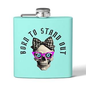 May include: A turquoise blue stainless steel hip flask with a silver lid and a skull wearing pink glasses and a black and white checkered bow. The text "Born to Stand Out" is printed in black around the skull.