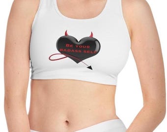 May include: White sports bra featuring a black heart design with red horns and a tail. The heart has the text "BE YOUR BADASS SELF" in red. The bra is a racerback style.