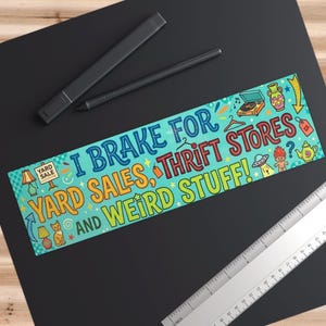 May include: A colorful banner with the text "I Brake for Yard Sales, Thrift Stores, and Weird Stuff!" is displayed on a black surface. The banner features illustrations of a record player, a UFO, and other quirky items. A ruler and two pens are also visible.