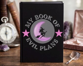 Puede incluir: Un libro negro con el texto "MY BOOK OF EVIL PLANS" en letras plateadas. La portada presenta una bruja en una escoba frente a una luna creciente, con estrellas rosas a cada lado. El libro está sobre una superficie de madera.
