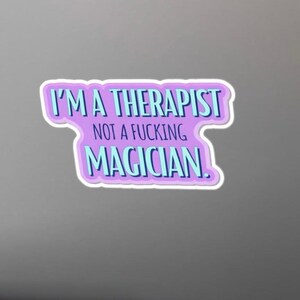 May include: A sticker with the text "I'M A THERAPIST NOT A FUCKING MAGICIAN" in blue and white lettering. The text is set against a purple background with a white border. The sticker is on a gray background.
