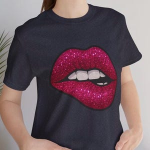 Puede incluir: Camiseta azul marino oscuro con un gran gráfico de labios magenta brillante. Los labios están ligeramente entreabiertos, revelando dientes blancos. El diseño está centrado en la camiseta, creando un look audaz y llamativo.