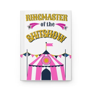 Könnte beinhalten: Ein weißes Notizbuch mit dem Text "RINGMASTER of the SHITSHOW" in goldfarbenen und blauen Buchstaben. Das Cover zeigt eine Illustration eines rosa-weiß gestreiften Zirkuszeltes mit goldenem Sternmuster.