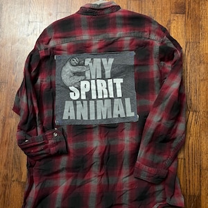 May include: A long-sleeve button-down shirt in a red and black plaid pattern. A large rectangular gray patch is sewn onto the back, featuring a graphic illustration of a sloth and the white text that reads MY SPIRIT ANIMAL in block letters.