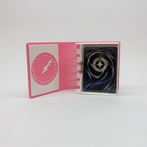 May include: A pink and white plastic life counter case featuring a circular dial with numbers zero through twenty and a white pointer. The case is open, revealing a rectangular slot containing a single dark playing card with a white star emblem.
