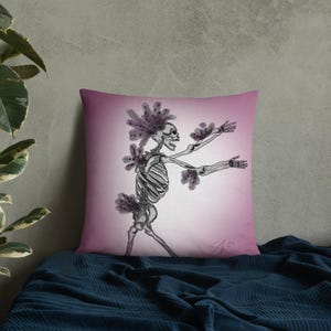May include: Decorative throw pillow with a skeleton illustration. The black and white skeleton is adorned with purple floral accents and is set against a pink gradient background. The pillow is square and rests on a blue surface.