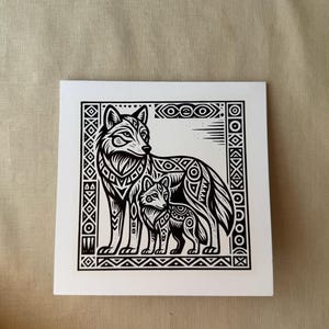以下が含まれることがあります： 大人と子供の2匹のオオカミの白黒イラスト。複雑な幾何学模様が描かれています。アートワークは装飾的なボーダーで額装され、ニュートラルな背景に配置されています。画像は正方形の白いカードにあります。