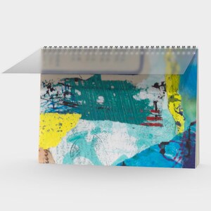 Könnte beinhalten: Ein Spiral-Schreibtischkalender mit einer transparenten Kunststoffabdeckung. Die Kalenderseiten zeigen ein abstraktes Kunstdesign mit leuchtenden Farben, darunter Türkis, Gelb, Blau und Rot. Das Kunstwerk scheint eine Mischung aus Pinselstrichen und Texturen zu sein.