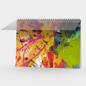 May include: A desk calendar with a spiral binding, showcasing abstract art. The artwork is a vibrant mix of yellow, pink, red, green, and blue paint splatters and streaks. The calendar is open, with a clear plastic sheet protecting the current month's artwork.