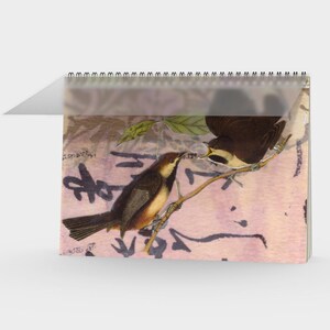 Könnte beinhalten: Ein Spiral-Kalender mit einer Vintage-Illustration von zwei Vögeln auf einem Ast. Die Vögel sind braun und weiß, vor einem rosa-beigen Hintergrund mit schriftartigem Text. Der Kalender ist offen, mit einer transparenten Kunststoffabdeckung.